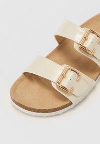 Beige sandaler med to glossy stropper, gyldne metalspænder, korklignende fodseng og hvid gummisål. Glat tekstur med flad design.
