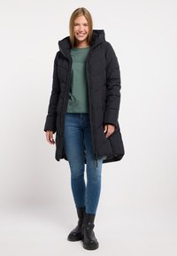 Black padded coat with a hood, zip closure, and long sleeves. Worn over a green top, paired with blue jeans and black ankle boots.