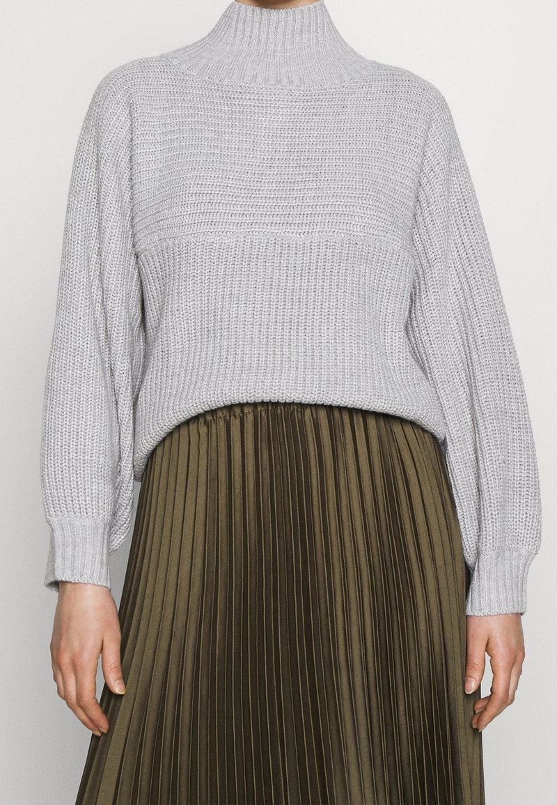 Pull léger en maille gris clair avec un col haut et des épaules tombantes, associé à une jupe plissée vert olive avec des lignes verticales et une texture douce.