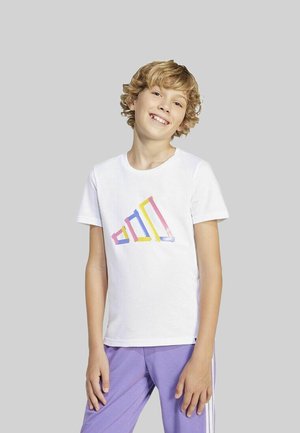 Lächelndes, blondes Kind mit lockigem Haar, das ein weißes T-Shirt mit buntem geometrischen Design und lila Hosen vor einem grauen Hintergrund trägt.