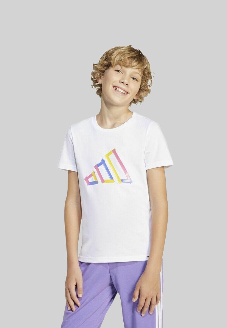Bambino biondo sorridente con capelli ricci che indossa una maglietta bianca con un design geometrico colorato e pantaloni viola davanti a uno sfondo grigio.