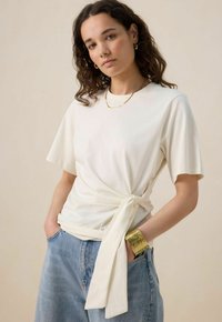 T-shirt ivoire à manches courtes avec un détail en nœud sur le côté, fabriqué en tissu doux. Assorti à un jean bleu clair, doté d'un fit décontracté.
