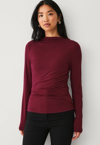 Bordeaux langemouw T-shirt met een getailleerde silhuet, zachte textuur en gerimpelde details bij de taille, gecombineerd met zwarte broek.