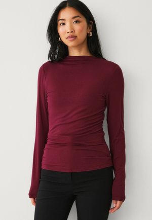 Blusa burdeos de manga larga con silueta ajustada, textura suave y detalles fruncidos en la cintura, combinada con pantalones negros.