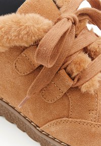 Stivale in suede marrone chiaro con bordo in pelliccia sintetica, lacci intrecciati e suola in gomma testurizzata. Include cuciture decorative e dettagli in morbido tessuto.