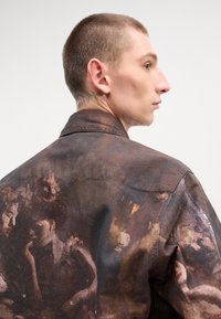 Chemise en tissu brun avec des imprimés artistiques et une apparence texturée. Col classique et design structuré visibles de côté.