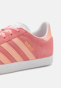 Rosa Wildleder Adidas Gazelle Sneaker mit Pfirsichstreifen, weißer Sohle und Schnürsenkeln, von der Seite und leicht schräg dargestellt.