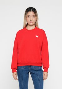 Sweatshirt rouge avec un col rond, des poignets et un ourlet côtelés, présentant un petit graphique en forme de cœur blanc sur la poitrine, fabriqué à partir d'un doux mélange de coton.