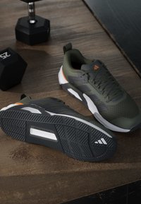 Paire de chaussures de sport Adidas vertes et noires posées sur un sol en bois à côté de deux haltères noirs de 25 livres.