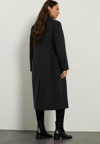 Kobieta z długimi brązowymi włosami, ubrana w czarny płaszcz do kolan, czarne spodnie i czarne botki na obcasie, stoi przy gładkiej ścianie.