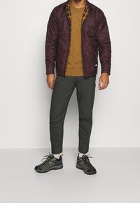 Prošívaná vínová bunda přes hořčicový svetr, spárovaná s úzkými tmavě zelenými kalhotami a černými turistickými botami s oranžovými akcenty.