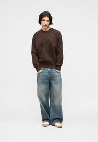 Νεαρό άτομο με σκουρόχρωμα μαλλιά που φοράει καφέ φούτερ, φαρδιά ξεθωριασμένα μπλε τζιν και λευκά αθλητικά παπούτσια, στέκεται με τα χέρια στις τσέπες.