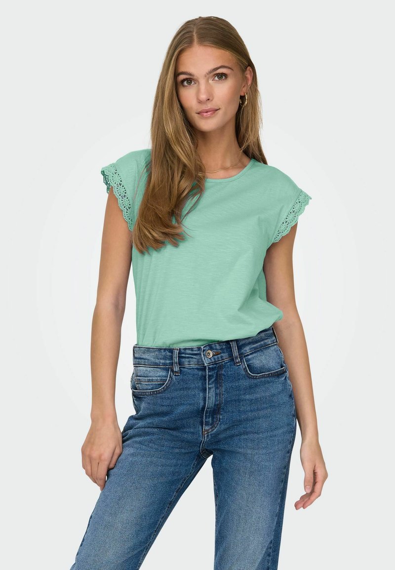 Mintgrünes, kurzärmliges T-Shirt mit Spitzenbesatz an den Ärmeln, kombiniert mit hochgeschnittenen blauen Jeans, die eine legere Passform zeigen.