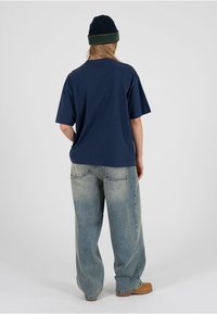 Camiseta de algodón azul marino de corte holgado, combinada con vaqueros de pierna ancha azul claro y botas marrones. La modelo lleva un gorro oscuro con acentos verdes.