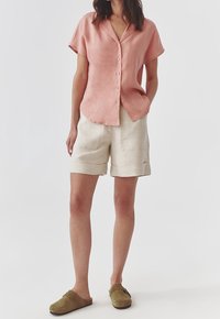 Camicetta in lino rosa chiaro a maniche corte con colletto, abbinata a pantaloni corti in lino beige con orli arrotolati e scarpe scivolate verde oliva.