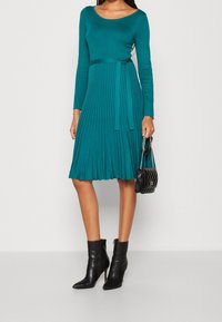 Vestito plissettato color verde petrolio con maniche lunghe e corpetto aderente, accentuato da una cintura in vita. Indossato con stivaletti neri alla caviglia e borsa trapuntata.