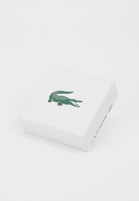 Bijela kartonska kutija s teksturiranom površinom, na vrhu s zelenim logom krokodila i natpisom "LACOSTE" sa strane.
