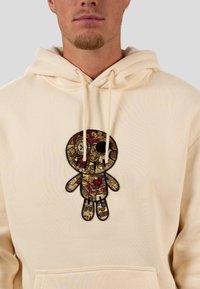 Light beige hoodie with a textured, embroidered doll design in gold, red, and black. Features drawstrings and a front pocket.