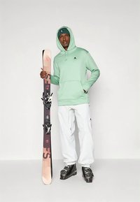 Mėtų žalios spalvos džemperis su kapišonu, baltos slidinėjimo kelnės, juodi slidinėjimo batai ir slides laikantis slidinėjimo lazda su rožiniu ir tamsiai violetiniu dizainu bei tvirtinimais. Paprastas ir funkcionalus aprangos komplektas.