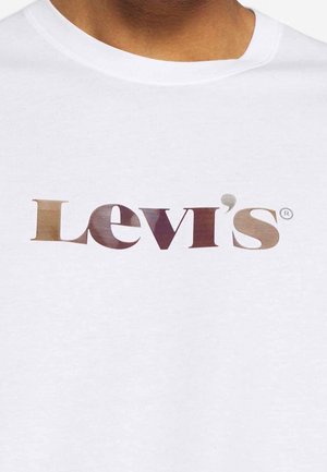 Biała koszulka noszona przez osobę, na której widoczna jest częściowo szyja i brązowa skóra, z dużym logo Levi's w brązowym gradientowym kolorze na klatce piersiowej.