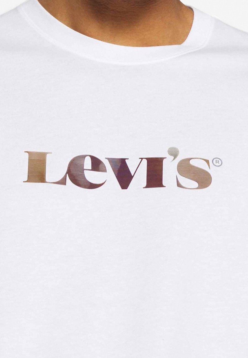 T-shirt blanc porté par une personne, avec une partie du cou visible et une peau marron, arborant le logo Levi's en grandes lettres dégradées marron sur la poitrine.