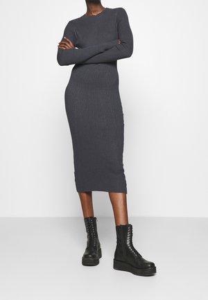 Zwarte midi-jurk met lange mouwen en geribbelde stof, gecombineerd met zwarte veterplateau laarzen, gedragen door een staand persoon met gekruiste armen.