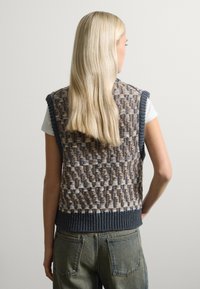 Chaleco de punto con un patrón tejido texturizado en tonos terrosos, con un dobladillo acanalado oscuro y mangas cortas, llevado sobre una camisa blanca.