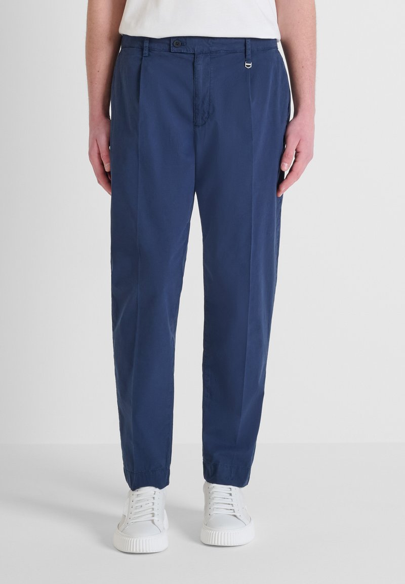 Pantaloni tapered blu navy realizzati in tessuto leggero, con pieghe, chiusura a bottone e polsini elasticizzati. Indossati con sneakers bianche.