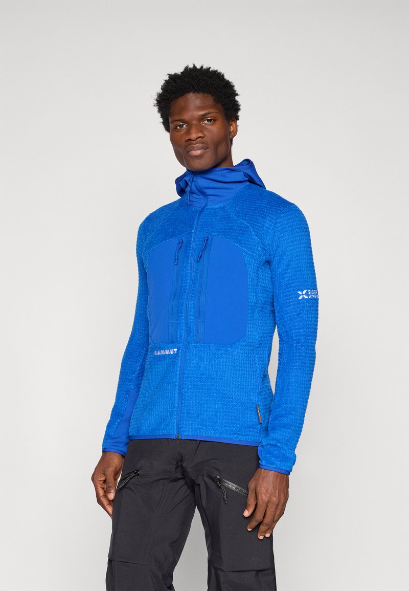 Μπλε, υφασματική ζακέτα με τσέπες με φερμουάρ, κουκούλα και αντίθετα λείες πάνελ; φορεμένη με μαύρο παντελόνι με τσέπες με φερμουάρ στα πλάγια.