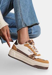 Les sneakers présentent une base en cuir blanc avec des accents en suède marron et un imprimé léopard. Semelle épaisse, collier en fourrure et lacets crème.