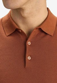 Brun texturerad pikétröja med ribbad krage, tvåknäppning och rundad nederkant. Materialet verkar vara stickad tyg.
