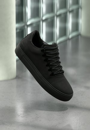 Mattschwarzer Sneaker mit dicker Sohle und schwarzen Schnürsenkeln, der über einem glatten grauen Boden schwebt, vor einer Glassteinwand und einem weißen Säulenhintergrund.