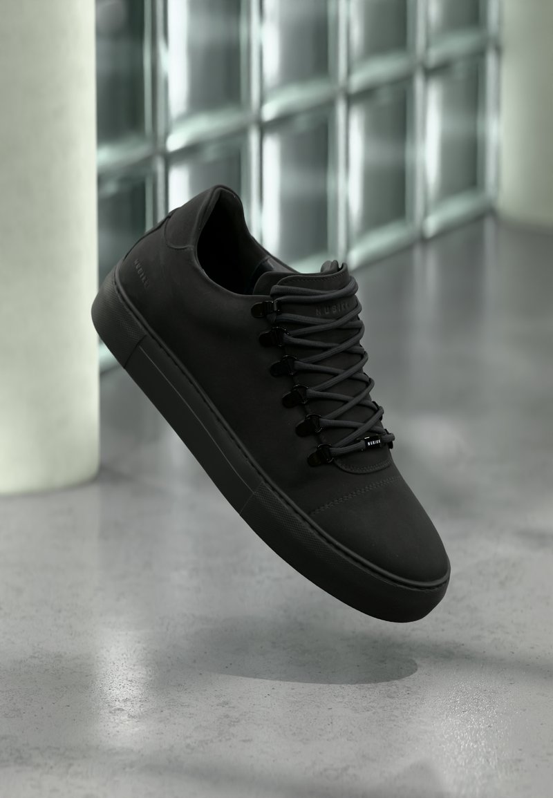 Matt svart joggesko med tykk såle og svarte lisser som svever over et glatt grått gulv, med glassblokkvegg og hvit kolonne i bakgrunnen.