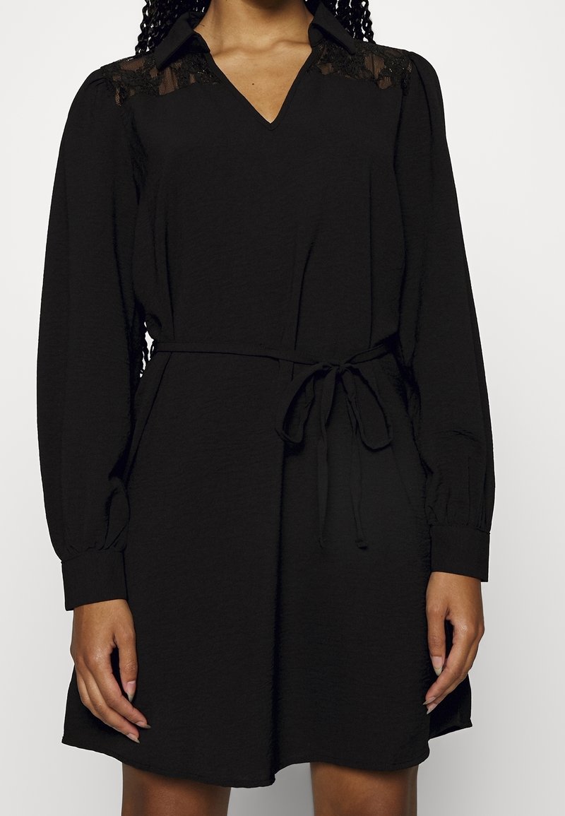 Robe noire à manches longues avec des détails en dentelle sur les épaules, un col en V et une ceinture nouée à la taille, portée par une personne aux cheveux tressés.