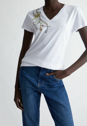 Osoba w białej koszulce z dekoltem w serek z wzorem złonej korony i łańcucha na piersi, w niebieskich dżinsach, z ręką w kieszeni.