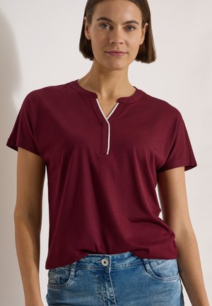 Femme portant un t-shirt bordeaux à manches courtes avec un détail de bordure blanche au niveau du col et un jean bleu en denim, debout devant un fond clair.