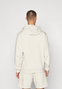 Felpa beige con cappuccio realizzata in un tessuto morbido, caratterizzata da una vestibilità ampia e polsini a coste. La vista posteriore mostra la semplicità del design e il colore neutro.
