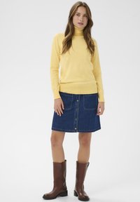 Pull col roulé jaune, jupe en denim bleu avec boutons sur le devant, bottes marron montantes jusqu'aux genoux, texture lisse, manches ajustées, deux poches avant sur la jupe.