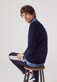 Marineblauer Pullover mit Rundhalsausschnitt über einem hellblauen Jeanshemd, kombiniert mit schwarzen Jogginghosen mit weißen Streifen und weißen Sneakers.