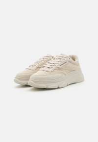 Beige suède sneakers hebben een dikke, gestructureerde zool, een ronde neus en een vetersluiting, met geperforeerde accenten en merknaam aan de zijkant.