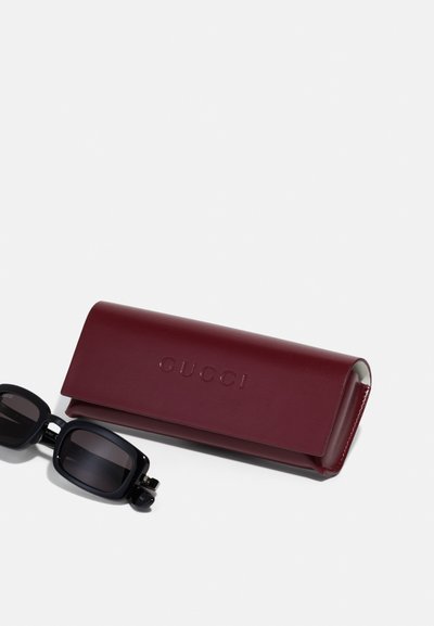 Lunettes de soleil noires avec une monture rectangulaire, accompagnées d'un étui en cuir bordeaux avec une marque en relief. Texture lisse et design épuré.