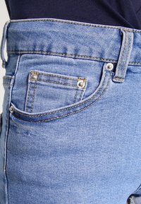 Vaaleansiniset farkut lähikuvassa, jossa on pieni etutasku, metalliset niitit ja erottuva tikkaus saumoissa.