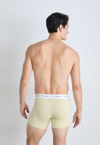Gele katoenen boxershorts met een witte elastische tailleband waarop "Calvin Klein" staat, van achteren gezien op een blote torso.