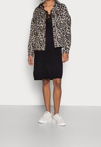 Giacca catena leopardata con chiusura a bottone, indossata su un vestito di maglia nera, abbinata a sneakers bianche. Palette di colori solidi e design testurizzato.