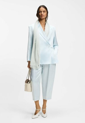 Frau trägt hellblauen maßgeschneiderten Anzug mit kurzen Hosen, weißem Schal, weißen High Heels und hält eine beige Handtasche, steht vor weißem Hintergrund.