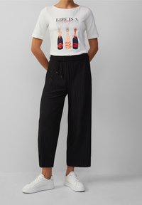 Witte t-shirt met een kleurrijke champagneflesafbeelding, gecombineerd met zwarte geplooide wijde broek en witte sneakers.