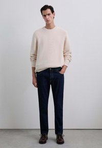 Beige gebreide trui, donkerblauwe jeans, bruine veterschoenen. Model staat tegen een neutrale achtergrond met de handen in de zakken. Eenvoudig, modern ontwerp.