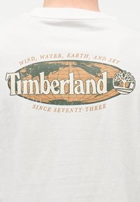 Λευκό πίσω μέρος πουκάμισου με ξεθωριασμένο λογότυπο Timberland, ένα στυλιζαρισμένο παγκόσμιο χάρτη, έμβλημα δέντρου και το κείμενο "Ανέμος, Νερό, Γη και Ουρανός από το εβδομήντα τρία."