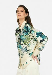 Blumenbluse aus Seide mit einer cremefarbenen Basis. Sie besitzt übergroße Blumenmuster in Blau- und Grüntönen. Mit Knopfkragen und langen Ärmeln.