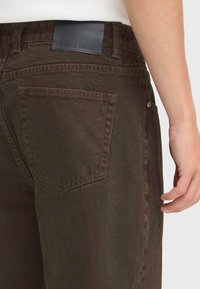 Mørkebrune denimjeans med en baklomme, etikett på topp linning som leser "CASUAL A VAGUE," og synlig søm. Stoffet har en glatt tekstur.